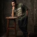 Man wearing a green t-shirt with text, leaning against a stone wall with an open book on a stool.